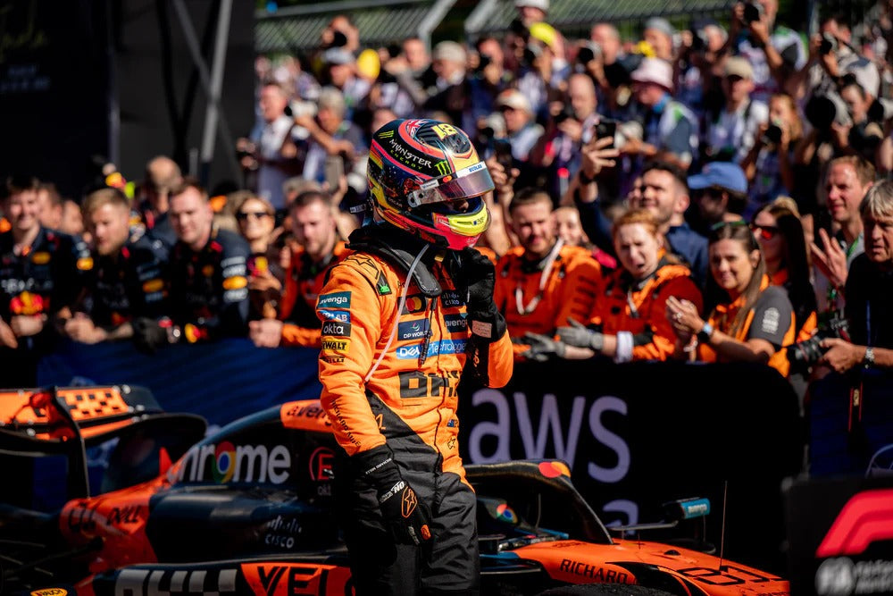Oscar Piastri in orange 2025 McLaren F1 racing suit and helmet standing next to a Formula 1 car with AWS branding, surrounded by a crowd - Kartx Gear